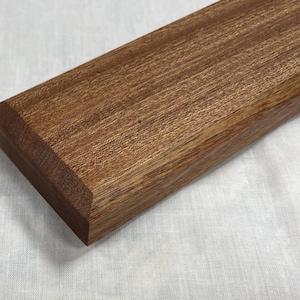 May include: A rectangular wooden box with a rich brown color and visible wood grain. The box has a beveled edge on one end and is resting on a white fabric surface. The box appears to be handcrafted.
