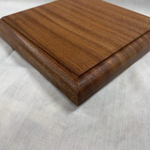 Puede incluir: Una tabla de cortar cuadrada de madera con bordes redondeados. La tabla está hecha de madera de color marrón rojizo y tiene una superficie lisa y pulida. Los bordes son ligeramente curvos, agregando un toque de elegancia al diseño.