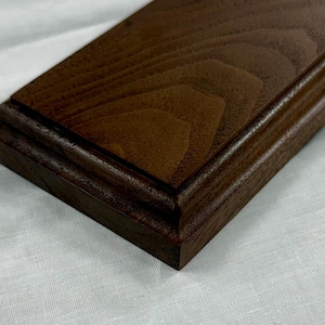 May include: A rectangular wooden display base featuring a dark brown finish with a visible natural wood grain pattern. The block has a bevelled edge profile that creates a stepped design along the perimeter, providing a flat surface for mounting.