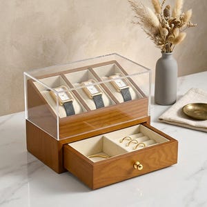 May include: A wooden watch and jewelry box with a clear acrylic lid. The top section displays three watches with gold-toned cases and gray bands. The bottom drawer holds rings and a bracelet. The box is on a marble surface.