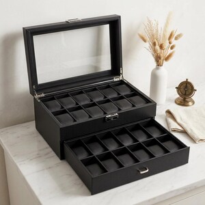 May include: A black watch box with a clear glass top and a pull-out drawer. The box has multiple compartments for storing watches. The box is made of a textured material and has a silver clasp and drawer pull.