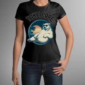 Puede incluir: Camiseta negra con un diseño gráfico de un oso polar con gafas de sol, frente a una puesta de sol con palmeras. El texto "TAKE CARE" está en un diseño circular. La camiseta la lleva una persona.