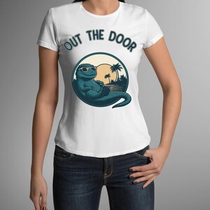 Puede incluir: Camiseta blanca con un dinosaurio relajado con gafas de sol, sobre un fondo de puesta de sol. El texto "OUT THE DOOR" está arqueado sobre el gráfico. La camiseta se combina con vaqueros azules.