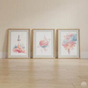 Könnte beinhalten: Drei gerahmte Aquarelldrucke. Die Drucke zeigen eine Rakete, einen Planeten mit Ringen und einen Astronauten in einer Wolke aus Farbe. Die Rahmen sind aus hellem Holz und die Kunst hat ein Weltraumthema.
