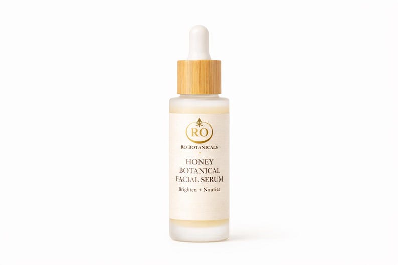 Pu&ograve; includere: Una bottiglia di vetro smerigliato di Honey Botanical Facial Serum con tappo e contagocce in legno. L'etichetta presenta testo dorato e un logo. Il siero &egrave; progettato per illuminare e nutrire la pelle.