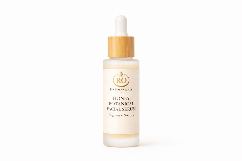 Pu&ograve; includere: Flacone in vetro smerigliato di Honey Botanical Facial Serum con tappo e contagocce in legno. L'etichetta presenta testo dorato e un logo. Il siero &egrave; progettato per illuminare e nutrire la pelle.