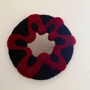 Op de afbeelding: Een ronde wandspiegel met een dik, getuft frame met een golvend, organisch patroon. Het frame is gemaakt van zacht, getextureerd garen in afwisselende segmenten van diepzwart en felrood, rondom een centraal reflecterend glazen oppervlak.