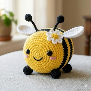 Pode incluir: Um brinquedo de abelha de crochê, com listras amarelas e pretas, asas brancas e uma pequena flor. A abelha tem olhos pretos, antenas e pequenos pés pretos. A abelha está sorrindo e tem bochechas rosadas.
