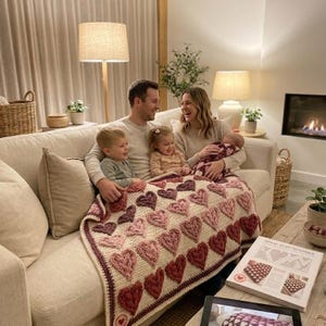 Könnte beinhalten: Eine fünfköpfige Familie sitzt auf einem cremefarbenen Sofa, bedeckt mit einer handgefertigten Häkeldecke. Die Decke hat ein sich wiederholendes Herzmuster in Rosa- und Burgundertönen. Ein Buch mit Häkelanleitungen liegt auf dem Couchtisch.