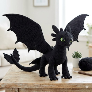 Puede incluir: Un dragón negro de ganchillo con grandes alas, una larga cola y ojos verdes. El dragón hecho a mano está posado sobre una mesa de madera. Una madeja de hilo negro, un ganchillo y una pequeña planta en maceta también están sobre la mesa.