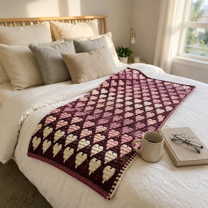 Könnte beinhalten: Handgefertigte Häkeldecke mit Herzmuster in Burgunder-, Rosa- und Cremetönen. Die Decke liegt auf einem Bett mit Kissen und einem Buch. Das Design der Decke zeigt Reihen von Herzen.
