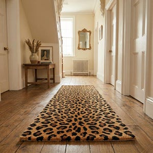 May include: A rectangular leopard print rug in a hallway. The rug features a tan background with dark brown spots. The rug is placed on a wooden floor, and the hallway has white doors and walls.