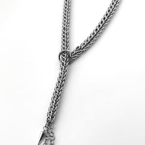 Könnte beinhalten: Eine silberfarbene Kettenhalskette mit einem geflochtenen Metallglieddesign. Die Halskette verfügt über einen kreisförmigen Ringverbinder, der einen herabhängenden Kettenabschnitt hält, welcher in drei spitzen Metallkegeln am unteren Ende endet.