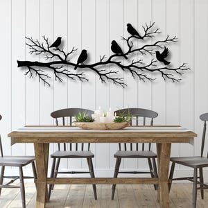 Op de afbeelding: Zwart metalen wandkunstwerk met een boomtak met verschillende vogels. De vogels zijn silhouetten tegen een witte muur. Het kunstwerk bevindt zich boven een houten eettafel met stoelen.