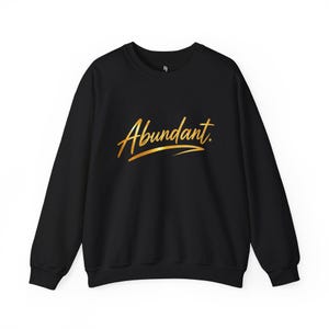 Könnte beinhalten: Ein schwarzes Langarm-Sweatshirt mit Rundhalsausschnitt, auf dessen Brust das Wort Abundant in goldfarbener Schreibschrift steht. Der Schriftzug enthält einen horizontalen Unterstrich unter dem Wort und ein kleines Logo am Halsausschnitt.