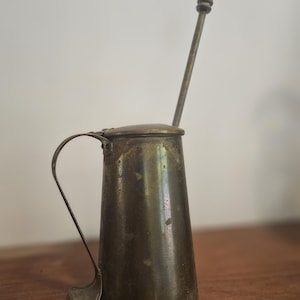 Op de afbeelding: Een vintage messing kan met een lange handgreep en een deksel. Een lange, dunne roerstaaf steekt uit de kan. De kan heeft een licht conische vorm en een aangetaste afwerking, wat duidt op leeftijd en gebruik. De handgreep is gebogen en aan de zijkant bevestigd.