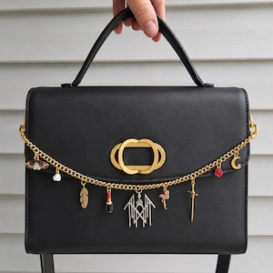 May include: A black structured handbag featuring a gold-toned oval clasp and a decorative gold chain draped across the front. The chain is adorned with various charms, including a moth, feather, lipstick, silver emblem, flamingo, sword, rose, and moon.