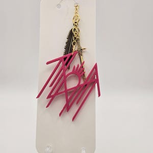 Peut inclure: Un pendentif rose avec un motif géométrique et le mot "TAH" est attaché à une chaîne dorée avec un charme de plume et d'épée. Le pendentif est présenté sur une carte blanche avec un trou en haut.
