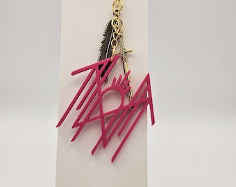 May include: A pink pendant with a geometric design and the word "TAH" is attached to a gold chain with a feather and sword charm. The pendant is displayed on a white card with a hole at the top.