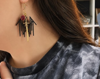 May include: A gold-toned earring featuring a dangling gold sword charm, a small red rose accent, and a black geometric, angular pendant. The earring is attached to a gold-toned hook wire, creating a layered, multi-element design that hangs vertically.