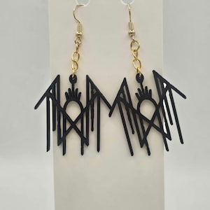 May include: A pair of black geometric dangle earrings. The earrings feature a stylized design with a central sun-like shape and vertical lines, suspended from gold-colored ear wires and chain links. The earrings are displayed against a white background.