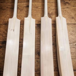 May include: Four light-colored wooden cricket bats arranged side by side. Each bat features a long, smooth blade with a visible wood grain and a slender handle wrapped in white grip tape. The bats have a classic shape with a distinct shoulder.