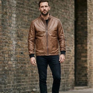 Könnte beinhalten: Eine braune Lederjacke mit Stehkragen, einem silbernen Reißverschluss in der Mitte und schwarzen Rippbündchen. Die Jacke wird über einem schwarzen Oberteil getragen und mit einer dunkelblauen Jeans kombiniert, was einen strukturierten Look ergibt.
