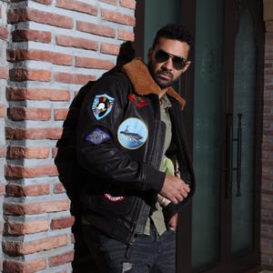 May include: Black leather jacket with multiple patches, a brown fur collar, and a zipper closure. The jacket features a Top Gun theme with patches on the chest and sleeves. The person in the image is wearing sunglasses and a green shirt.