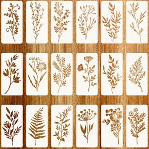 May include: A set of eighteen rectangular stencils featuring various botanical designs, including wildflowers, ferns, and leafy branches. Each stencil displays a unique plant silhouette in brown against a white background, arranged in a grid.