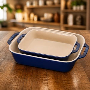 May include: Two nested, rectangular baking dishes. The outer dish is a deep blue, and the inner dish is a cream color. Both have handles and are sitting on a wooden surface. The background shows a kitchen setting.