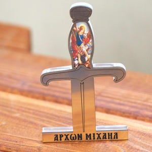 May include: A decorative metal sword with a detailed handle featuring a religious image. The sword has a silver blade and a rectangular base with Greek text. The handle has a white top.