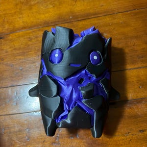 May include: A 3D printed figurine featuring a black outer shell with jagged, broken-looking segments that reveal a vibrant purple core underneath. The figure has two circular purple eyes and a small, simple horizontal line for a mouth.