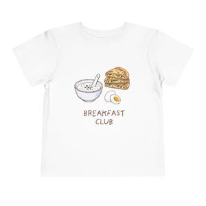 May include: White toddler t-shirt with a graphic of a bowl of cereal, a stack of bread, and a halved egg. The text "BREAKFAST CLUB" is printed below the graphic in brown. The shirt is made of a soft material.