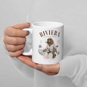 Könnte beinhalten: Weiße Keramik-Tasse mit einer Illustration eines Hundes auf einem Motorroller, der eine Zigarre raucht. Auf der Tasse stehen die Worte "RIVIERA STATE OF MIND" und "SAINT-TROPEZ", sowie eine Palme und eine Küstenlandschaft.