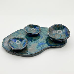 Op de afbeelding: Een set van drie keramische stukken met een grote, onregelmatig gevormde schaal met een glanzend, gevlekt blauw en groen glazuur. Twee kleine, bijpassende kommetjes met golvende, onregelmatige randen staan op de schaal en hebben hetzelfde blauw-groene kleurenpalet.