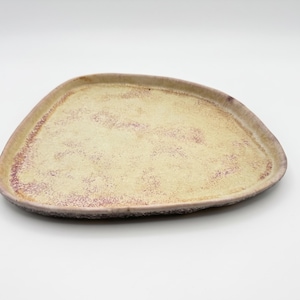 Op de afbeelding: Een onregelmatig, driehoekig keramisch bord met een getextureerd, lichtbeige oppervlak. De randen zijn voorzien van een gespikkeld, roodbruin glazuur dat overgaat in het crèmekleurige midden, wat een duidelijk contrast op het platte vlak creëert.