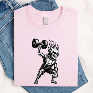 May include: A light pink t-shirt featuring a black and white graphic of a skeleton with flames for hair, wearing a hooded tank top and shorts, lifting a heavy dumbbell overhead with one arm while in a squatting position.