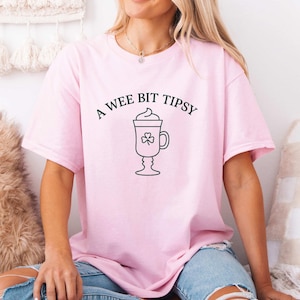 May include: A pale pink t-shirt featuring the phrase "A Wee Bit Tipsy" and a black line drawing of a drink with a shamrock. The shirt is a relaxed fit, suitable for casual wear or St. Patrick's Day.