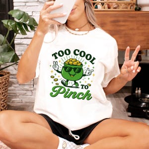 Puede incluir: Camiseta blanca con un gráfico del Día de San Patricio. El gráfico presenta una olla de oro verde con gafas de sol, con el texto "Too Cool to Pinch". La olla está rodeada de monedas de oro y tréboles.