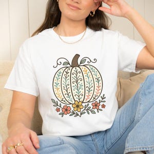 May include: White t-shirt featuring a detailed pumpkin design. The pumpkin is cream-colored with a brown stem and is decorated with green, yellow, and orange floral patterns. Small flowers in pink, yellow, and orange are at the base of the pumpkin.