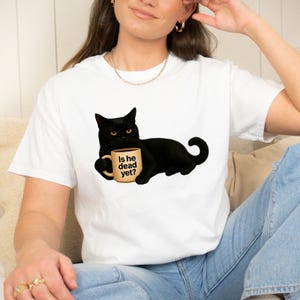 May include: A white short-sleeved t-shirt featuring a graphic of a black cat lying down and holding a tan mug. The mug has the text Is he dead yet? printed in black. The cat has yellow eyes and is positioned centrally on the front of the shirt.