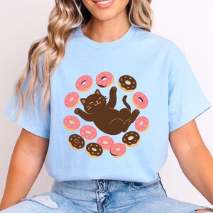 May include: A light blue t-shirt featuring a central graphic of a brown cat lying on its back, surrounded by a circular arrangement of pink and chocolate-colored donuts with sprinkles. The cat has a smiling expression and raised paws.