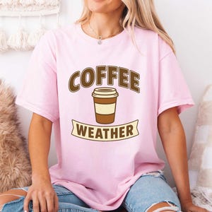 Może przedstawiać: Jasnoróżowa koszulka z napisem "COFFEE WEATHER" w brązowych literach i grafiką filiżanki kawy. Koszulka jest w stylu casual, idealna dla miłośników kawy.