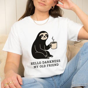 May include: A white short-sleeved t-shirt featuring a black graphic of a sloth holding a steaming mug. Below the sloth, the text reads "HELLO DARKNESS MY OLD FRIEND" in black capital letters. The shirt has a crew neckline and a standard fit.