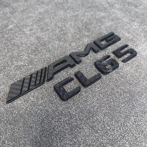 May include: A black automotive emblem featuring the text AMG CL65 with a carbon fiber pattern finish. The letters and numbers are raised with a glossy surface, displaying a distinct weave texture throughout the entire badge design.