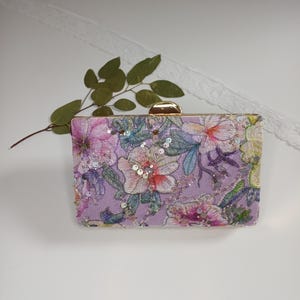 Può includere: Una pochette rettangolare con base in tessuto viola chiaro e un intricato ricamo floreale nei toni del rosa, verde e bianco. La borsa è impreziosita da piccole paillettes riflettenti e rifinita con una cornice e una chiusura in metallo dorato.