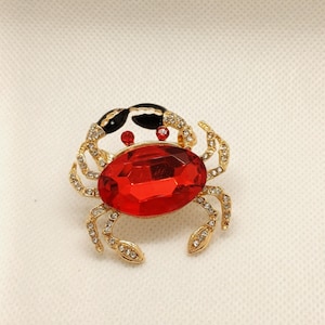 Peut inclure: Broche en forme de crabe doré avec un grand corps en pierre rouge facettée. Les yeux du crabe sont de petits gemmes rouges, et sa carapace est noire et dorée. Les pattes et les pinces sont ornées de petits cristaux transparents.