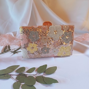 Pode incluir: Uma bolsa clutch retangular com um padrão floral composto por lantejoulas multicoloridas em tons de rosa, amarelo e verde sálvia. A bolsa possui um fecho metálico dourado no centro superior e uma silhueta estruturada e retangular.