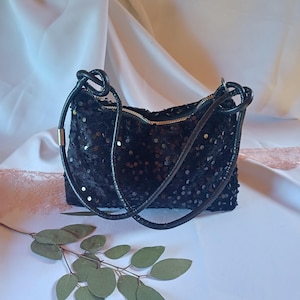 Pode incluir: Uma bolsa de mão preta coberta com pequenas lantejoulas refletoras. A bolsa possui um fecho de correr superior com um puxador metálico e duas alças de ombro pretas, finas e arredondadas, com nós nos pontos de fixação superiores.