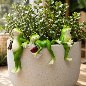 Puede incluir: Una maceta de cerámica gris claro con una exuberante planta verde, adornada con tres caprichosas figuras de ranas verdes. Una rana sostiene una botella, otra una copa de vino y la tercera un cóctel.